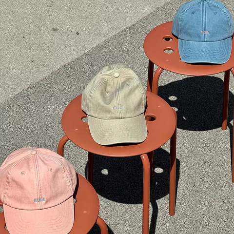Drei Caps liegen nebeneinander auf orangenen Stühlen. Die Caps sind in rosa mit der Aufschrift Gude, Khaki in Hello und Blau mit Moin