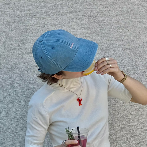 Blaue Cap mit der Aufschrift "Moin", getragen von einer weiblichen Person mit hellem T-Shirt, die in eine Zitrone beißt und dabei ein rotes Getränk in der Hand hält.